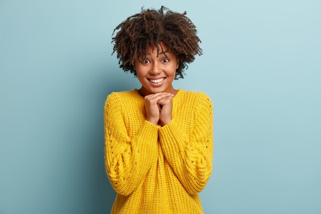 Afro-americana satisfeita tocada mantém as mãos juntas sob o queixo, sente admiração, expressa emoções positivas, tem um sorriso cheio de dentes, usa um macacão amarelo, se diverte e ri dentro de casa. Conceito de emoções