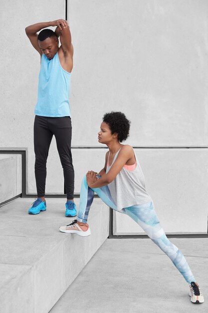 Afro-americano adulto masculino levanta as mãos, aquece antes do treinamento cardiovascular. Mulher de pele escura em leggings e tênis estica as pernas, se prepara para correr a maratona. Duas pessoas esportivas na escada