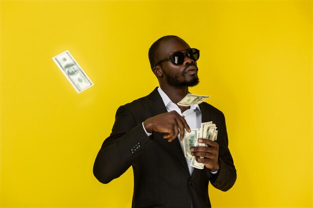 Afro-americano barbudo está jogando fora dólares de uma mão, usando óculos escuros e terno preto
