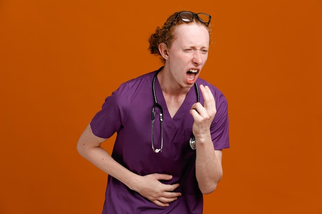 Agarrando estômago jovem médico masculino vestindo uniforme com estetoscópio isolado em fundo laranja