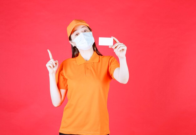 Agente de serviço feminino com dresscode cor laranja e máscara apresentando seu cartão de visita e apontando para outra pessoa
