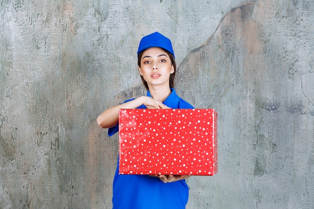 Agente de serviço feminino de uniforme azul, segurando uma caixa de presente vermelha com pontos brancos.