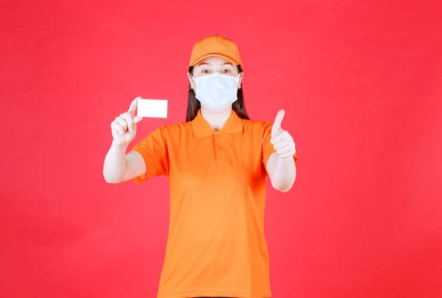 Agente de serviço feminino em dresscode cor laranja e máscara apresentando seu cartão de visita e mostrando sinal positivo com a mão