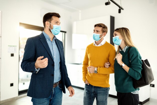 Agente masculino e jovem casal com máscaras faciais conversando enquanto procurava novos imóveis