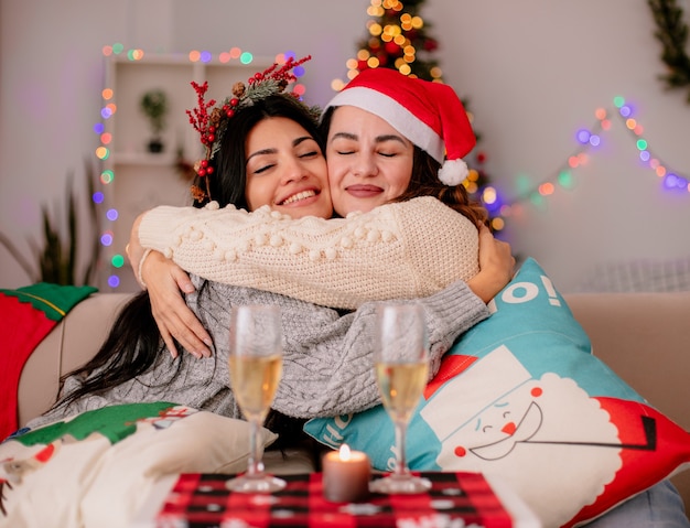 Foto grátis agradáveis garotas bonitas com chapéu de papai noel se abraçando sentadas em poltronas e curtindo o natal