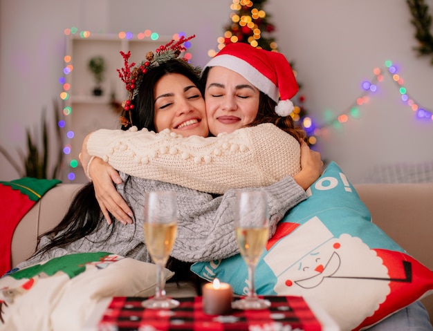 agradáveis garotas bonitas com chapéu de Papai Noel se abraçando sentadas em poltronas e curtindo o natal