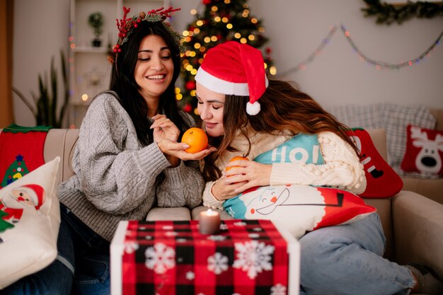 agradáveis garotas bonitas com chapéu de Papai Noel segurando e cheirando laranjas sentadas em poltronas e curtindo o natal em casa