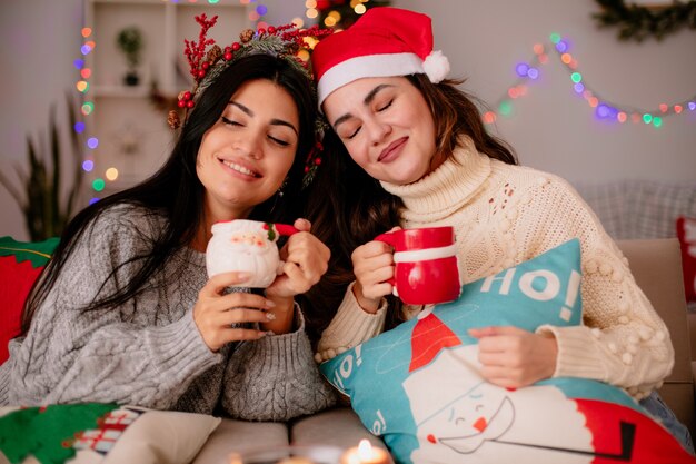 agradáveis garotas com chapéu de Papai Noel segurando copos sentados em poltronas e curtindo o Natal em casa
