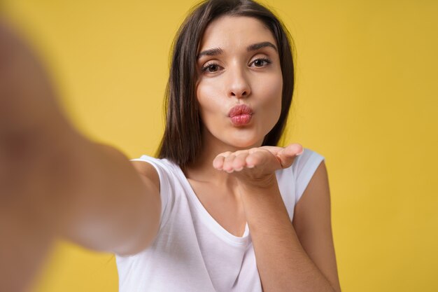 Agradável garota atraente fazendo selfie em estúdio e rindo. Mulher jovem bonita com cabelo castanho, tirando foto de si mesma em fundo amarelo brilhante.