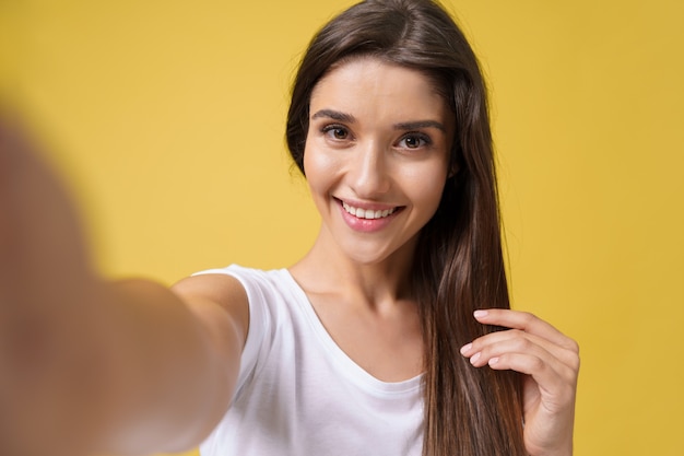 Agradável garota atraente fazendo selfie em estúdio e rindo. mulher jovem bonita com cabelo castanho, tirando foto de si mesma em fundo amarelo brilhante.