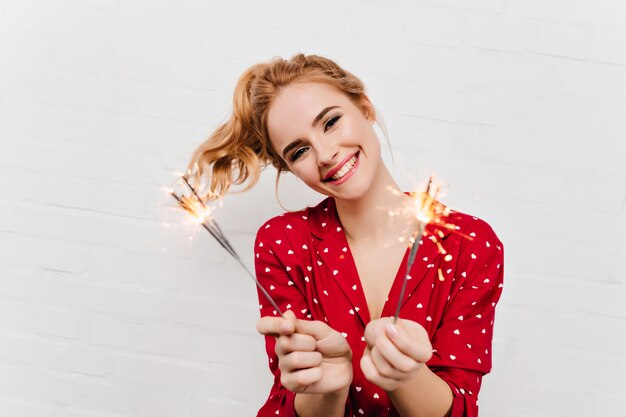 Agradável jovem vestida de vermelho segurando luzes de bengala. Foto interna de uma garota animada com cabelos loiros ondulados, comemorando o ano novo.