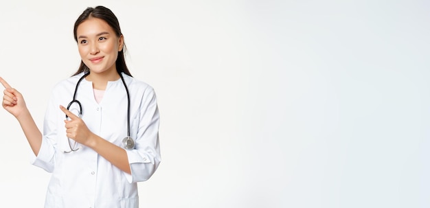 Agradável médica sorridente de uniforme apontando os dedos e olhando para a esquerda com um sorriso amigável e carinhoso