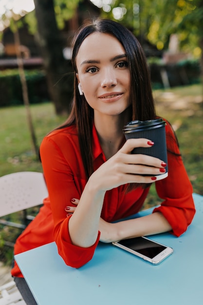 Agradável mulher morena de camisa vermelha se divertir no café de rua.