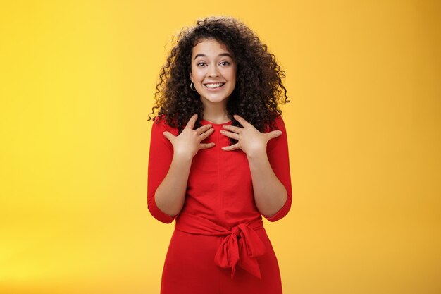 Agradecida e agradecida, terna menina gentil de cabelos cacheados em um vestido vermelho, apertando as mãos nos seios, sendo surpreendida com um presente terno e reconfortante inesperado, agradecendo, sorrindo encantada com o fundo amarelo