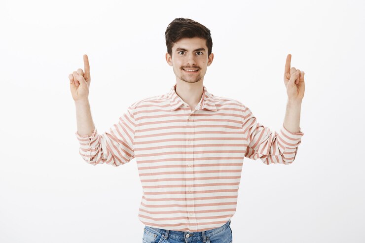 Foto grátis agradou um cara comum de aparência amigável e camisa listrada, levantando o dedo indicador e apontando para cima, sorrindo amplamente, sugerindo aos amigos que se sentassem no café no andar de cima, de pé