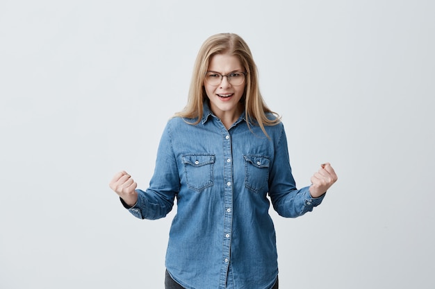 Agressão, reação humana negativa e atitude. Brava estressada mulher loira de óculos, camisa jeans gritando em voz alta com a boca aberta, punhos cerrados, tendo olhar louco e furioso