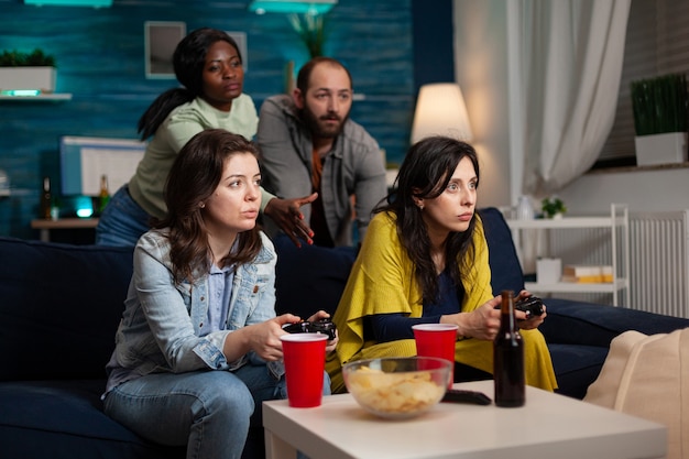 Agrupe amigos multiétnicos jogando videogame usando o controle durante a competição de batalha online. Pessoas se divertindo, bebendo cerveja, socializando tarde da noite enquanto estão sentadas no sofá da sala.