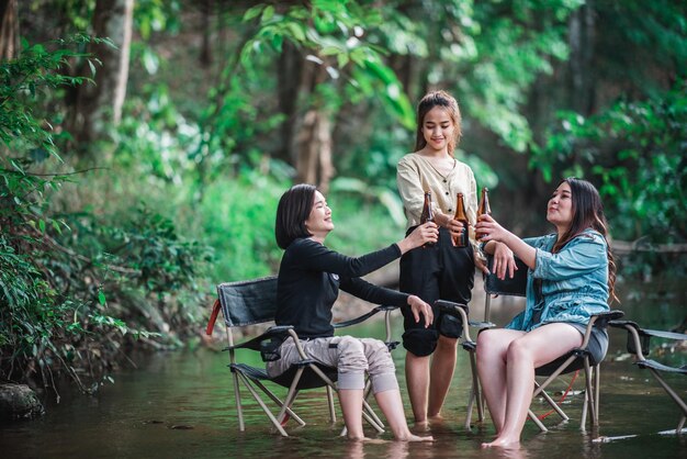 Agrupe belas mulheres asiáticas amigas viajantes relaxando em cadeiras de acampamento no córrego Elas estão torcendo e bebendo cerveja durante o acampamento conversando com diversão e felizes juntos