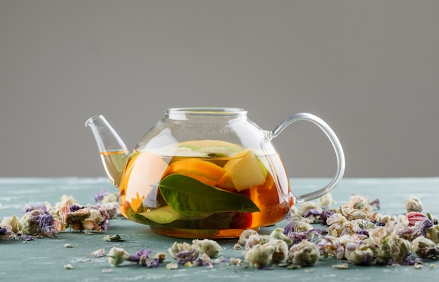 Água com infusão de frutas em um bule com vista lateral para flores secas na superfície de gesso e cinza