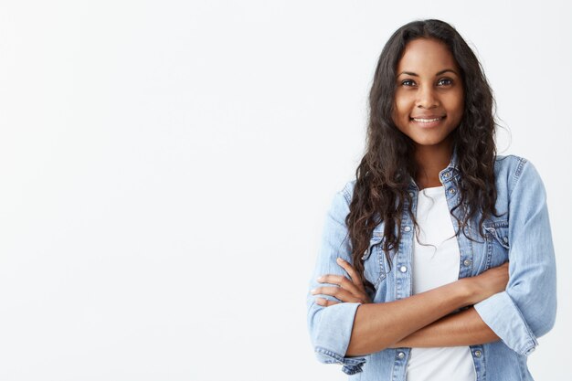 Ainda bem que a jovem mulher afro-americana bonita com pele escura e cabelos longos pretos posando dentro de casa com os braços cruzados, sorrindo amplamente com dentes brancos, rindo de uma boa piada, vestindo camisa jeans