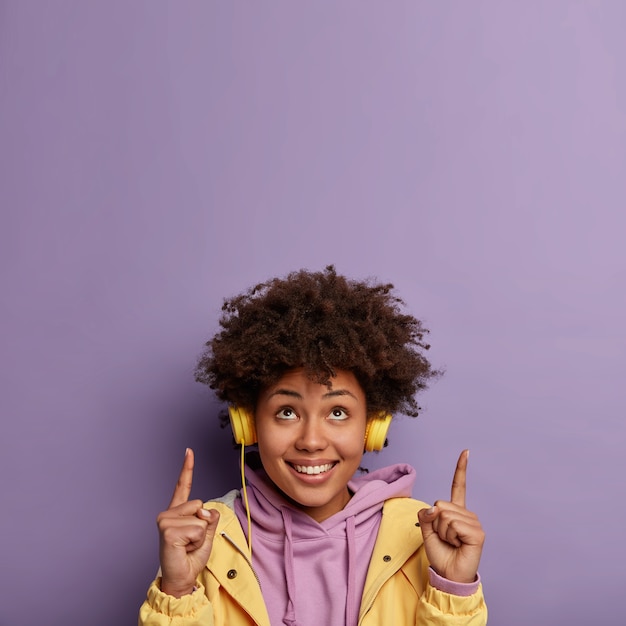 Foto grátis ainda bem que a mulher tem cabelo encaracolado, aponta os dois dedos para o teto, mostra o espaço da cópia acima, tem bom humor enquanto ouve canções divertidas