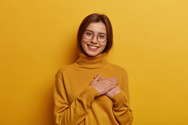 Foto grátis ainda bem que a rapariga europeia se sente satisfeita e grata, agradecida pelo presente maravilhoso, aperta as mãos no peito, tem um sorriso terno e agradável