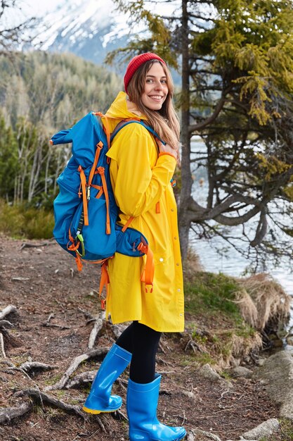 Ainda bem que jovem modelo feminina com mochila usa capacete vermelho, capa de chuva amarela e botas de borracha azuis