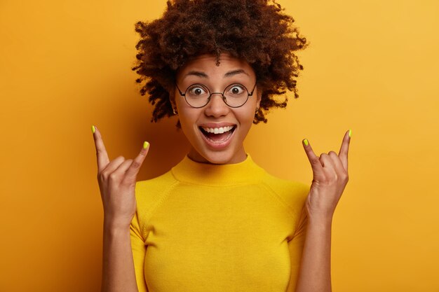 Ainda bem que mulher de pele escura faz sinal de heavy metal rock, sorri positivamente, ouve o gênero favorito de música, usa camiseta amarela casual e posa em ambientes internos. Rock n roll ao nosso redor. Conceito de linguagem corporal