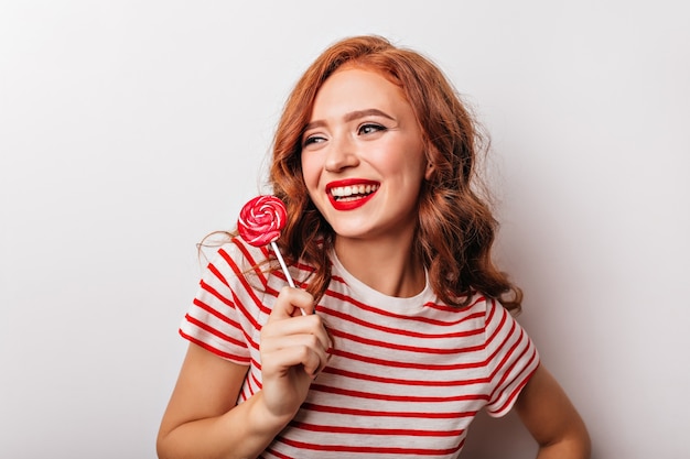 Foto grátis ainda bem que mulher ruiva segurando pirulito e rindo. glamourosa garota caucasiana com doces, aproveitando a sessão de fotos.