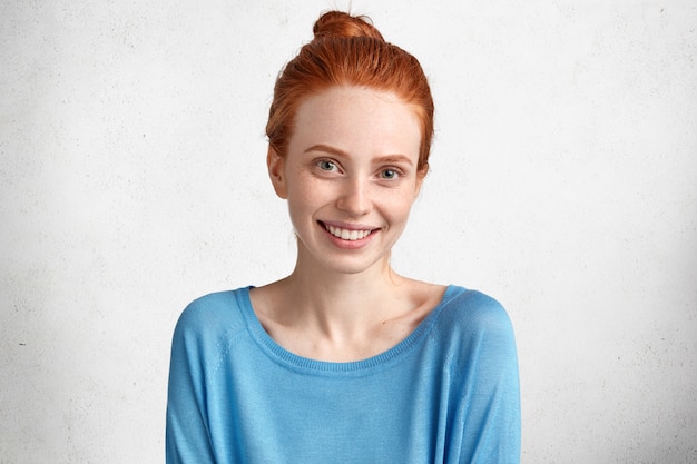 Ainda bem que mulher sorridente com pele sardenta e sorriso positivo, vestida com um sweater azul casual, estando em alto astral depois de um encontro com o namorado, isolada sobre o branco