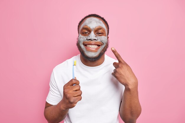 Ainda bem que o homem afro-americano sorri e se preocupa com os dentes segura escova de dentes e aplica máscara de argila de beleza no rosto