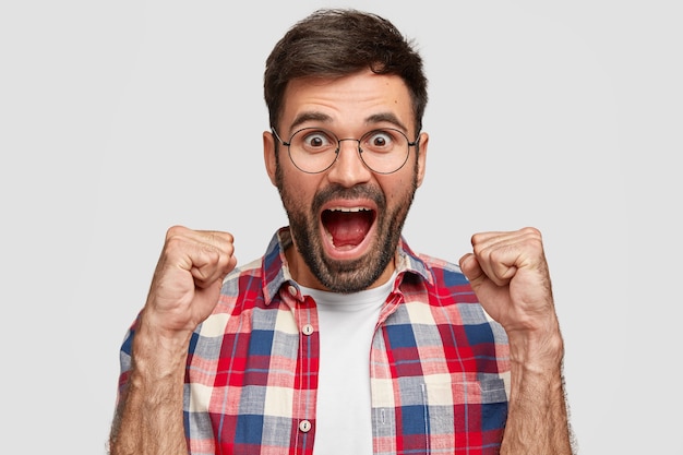 Ainda bem que o jovem feliz abre a boca, fecha os punhos e exclama em triunfo, vestido com uma camisa xadrez, de pé contra a parede branca. homem com barba por fazer se sente campeão ou vencedor