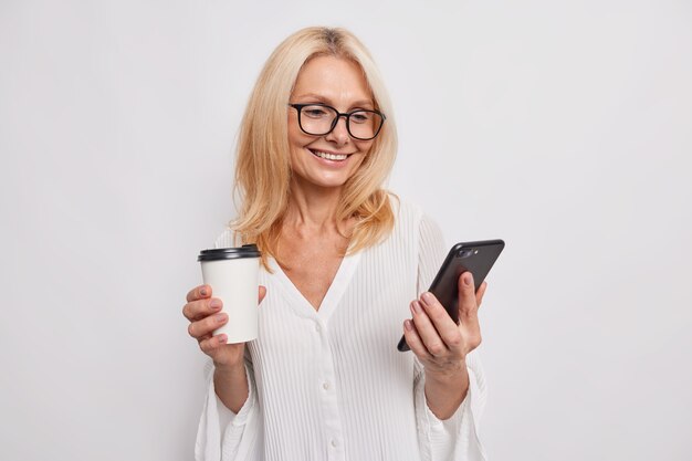 Ainda bem que uma mulher europeia bebe café num copo take-away segura no smartphone e utiliza ligação gratuita à Internet durante o intervalo, sorrisos, usa óculos e uma blusa elegante isolada sobre a parede branca