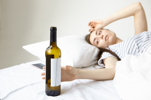 Alcoolismo e bebida. Mulher estressada na cama, sofrendo de uma terrível dor de cabeça depois da festa do álcool com amigos na noite passada, pegando uma garrafa de vidro de vinho, na esperança de aliviar a ressaca. Efeito filme