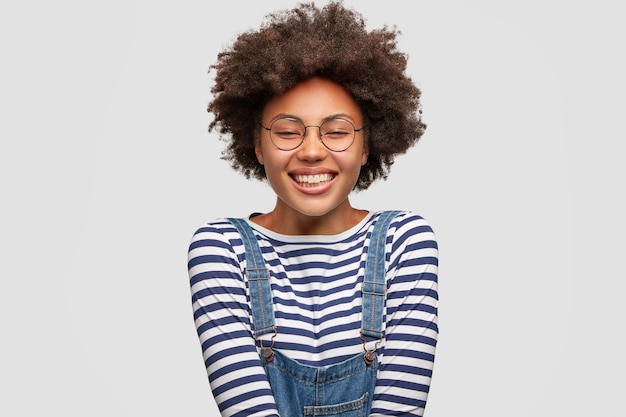 Alegre adorável mulher de pele escura com expressão satisfeita, tem sorriso largo, fecha os olhos de felicidade, veste um macacão da moda, expressa emoções positivas, isolada sobre uma parede branca.