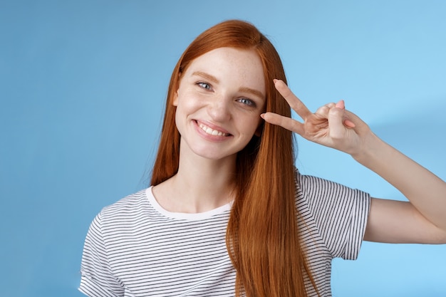 Alegre amigável linda ruiva olhando alegremente para mostrar o sinal da vitória da paz inclinando a cabeça fofos sorrindo amplamente os dentes brancos se divertindo, expressando otimismo de positividade, fundo azul de pé.