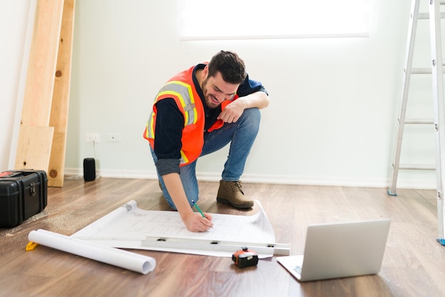 Alegre arquiteto latino sorrindo enquanto observa informações on-line no laptop e trabalha nos planos de construção