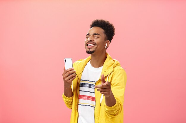 Alegre bonito afro-americano com penteado afro e barba fechando os olhos e sorrindo com prazer, curtindo música em fones de ouvido sem fio legais segurando um smartphone apontando para a câmera