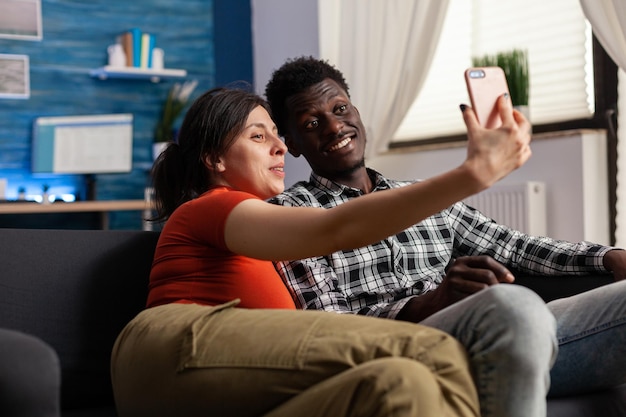 Alegre casal multiétnico posando para selfie sentado no sofá em casa. Pessoas adultas felizes casuais se unindo enquanto tiram selfies para se divertir usando o dispositivo smartphone na sala de estar