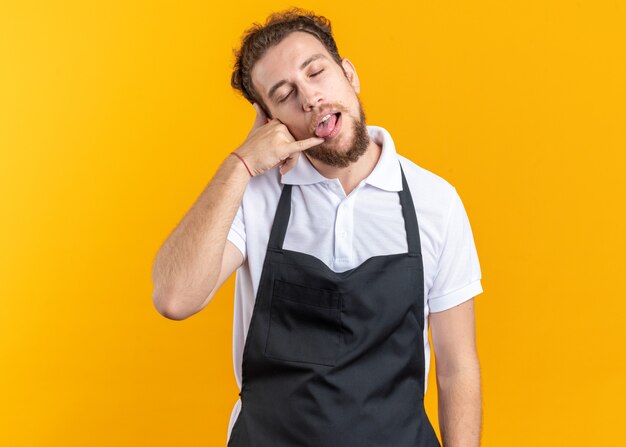 Alegre, com olhos fechados, jovem barbeiro masculino vestindo uniforme, mostrando gesto de língua e telefonema isolado na parede amarela