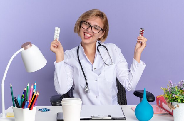 Alegre, com olhos fechados, jovem médica vestindo túnica médica com estetoscópio e óculos, sentada à mesa com instrumentos médicos segurando comprimidos e mostrando a língua isolada na parede azul