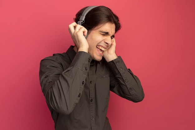 Alegre, com os olhos fechados, jovem bonito vestindo uma camiseta preta com fones de ouvido ouve música isolada na parede rosa