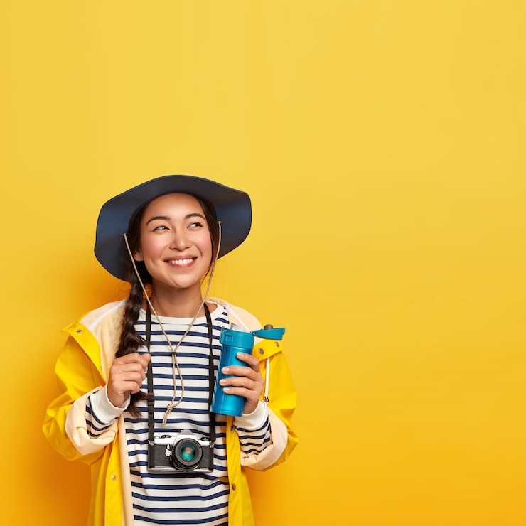 Foto grátis alegre e descontraída viajante feminina faz uma pausa para o café durante a longa jornada, explora a natureza, segura uma garrafa térmica com bebida, usa câmera retro, macacão listrado e capa de chuva
