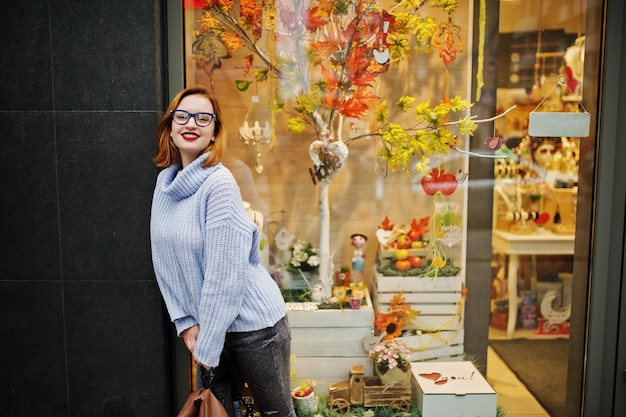 Alegre e linda mulher ruiva de óculos, suéter de lã azul quente com mochila posada ao ar livre contra folhas de outono decorações de árvores na loja