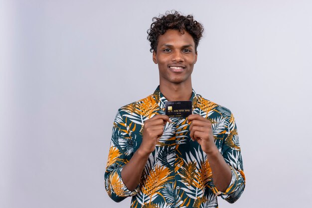 Foto grátis alegre e sorridente jovem bonito de pele escura com cabelo encaracolado, camisa estampada de folhas segurando e mostrando cartão de crédito