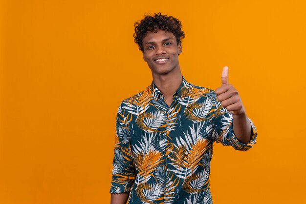 Alegre e sorridente jovem bonito de pele escura com cabelo encaracolado numa camisa estampada de folhas mostrando os polegares para cima