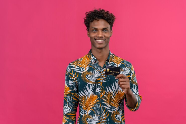 Foto grátis alegre e sorridente jovem bonito de pele escura com cabelo encaracolado numa camisa estampada de folhas, segurando e mostrando o cartão de crédito