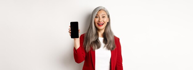 Alegre empresária asiática mostrando a tela do celular sorrindo e se divertindo sobre ba branca