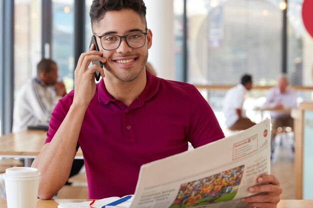 Alegre empresário com expressão alegre, usa óculos ópticos, conversa ao telefone, lê jornal, discute notícias com amigo, toma café em restaurante aconchegante. Freelancer profissional