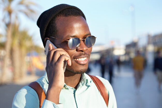 Alegre estudante afro-americano moderno na moda em óculos de sol redondos e chapéus fazendo ligação